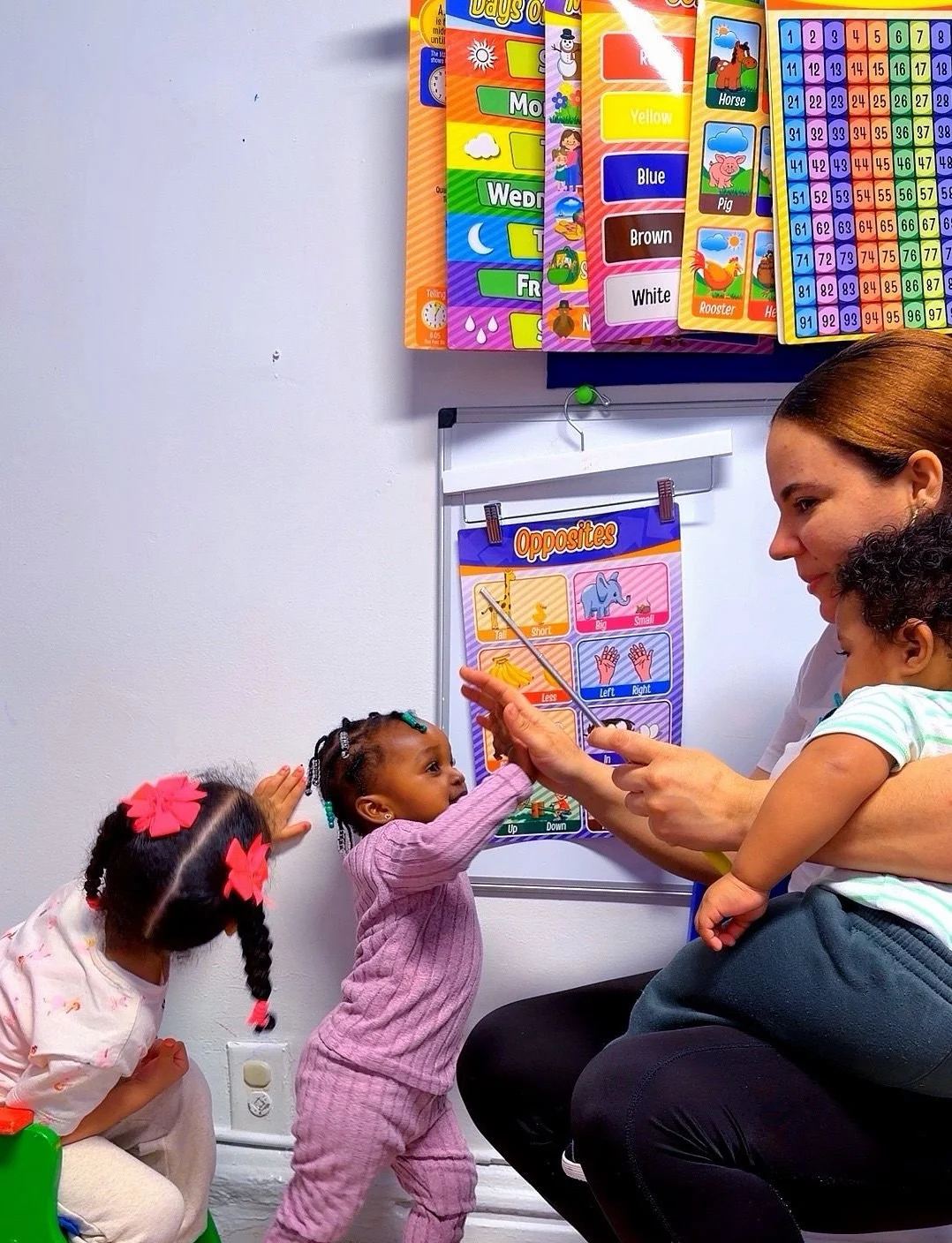 Teacher guiding three toddlers at the learning wall while holding a baby